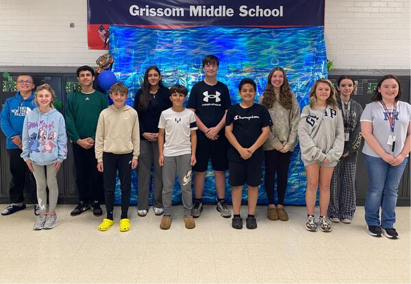 A group of students stands in front of lockers with a banner that reads "Welcome to Grissom Middle School Home of the Patriots".