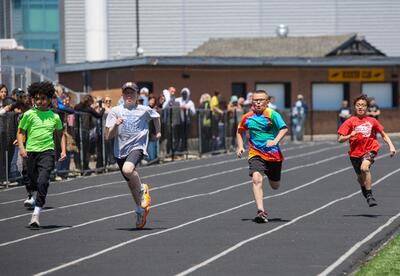 Four children race on a track, each in their own lane, with spectators watching from the sidelines.