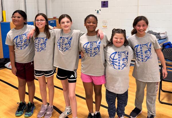 A group of six young people stand side-by-side, all wearing matching gray t-shirts with a blue cougar and basketball logo in school gymnasium.