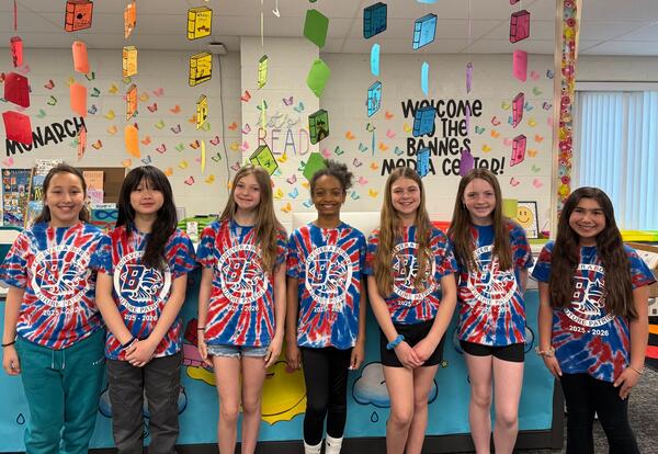 Six students wearing tye dye shirts stand in front in circulation desk in media center