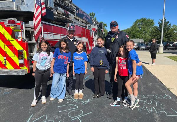 Group of children posing with two firefighters in front of a fire truck on a sunny day.