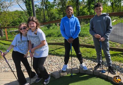 Four students stand and pose on a mini golf course holding putters.