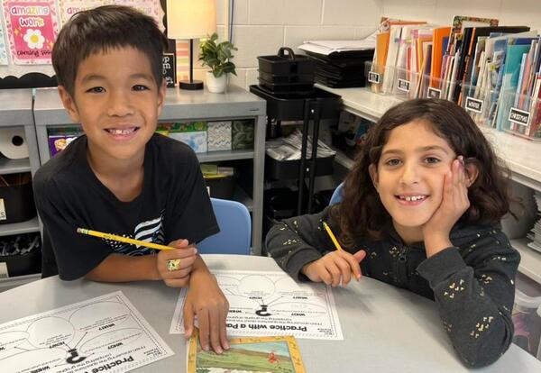 Two elementary students smile while working on activity at table in classroom.