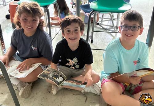 Three students smile while holding books sitting on the floor in classroom