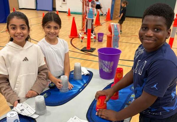 Three children smiling while stacking plastic cups on a table in a gymnasium setting.