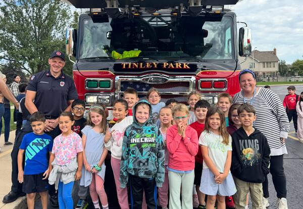 Group of students pose in rows with firefighter and teacher in front of fire engine outside