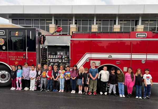 A group of children stand in a line in front of a red fire truck outside a brick building, posing for a group photo.