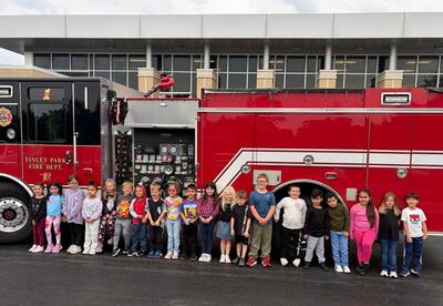 A group of children stand in a line in front of a red fire truck outside a brick building, posing for a group photo.