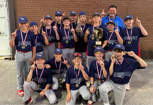 Softball team and coach pose outdoors in front of a brick wall, with several players holding medals and trophies, holding one finger up signifying 1st place