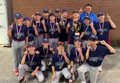 Softball team and coach pose outdoors in front of a brick wall, with several players holding medals and trophies, holding one finger up signifying 1st place