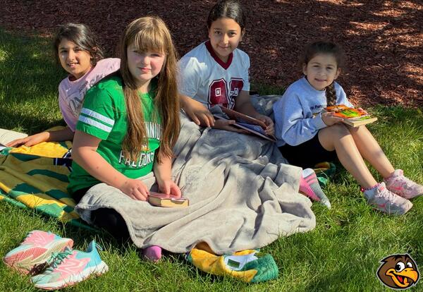 Four students are sitting on a colorful blanket on a grassy lawn, reading outside.