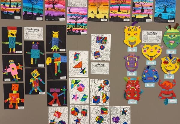 A wall display of colorful artwork: small landscape paintings in rows, many cartoon character drawings on dark and white backgrounds, and several bright animal masks with labels arranged across a beige wall.