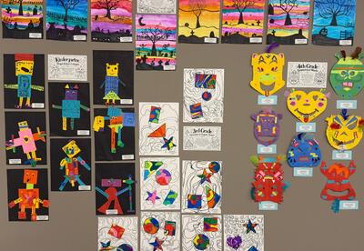 A wall display of colorful artwork: small landscape paintings in rows, many cartoon character drawings on dark and white backgrounds, and several bright animal masks with labels arranged across a beige wall.