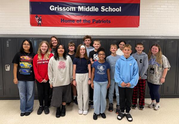 Students and staff of the month pose proudly in front of lockers in school hallway under a red banner that reads Welcome to Grissom Middle School - Home of the Patriots