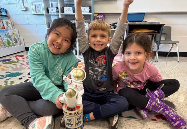 Three children sitting on classroom floor, smiling with arms raised, posing beside a small astronaut figure holding a canister.