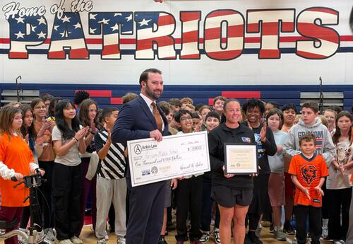 Person in suit holding a big check standing next to teacher holding a certificate in school gym in front of staff and students