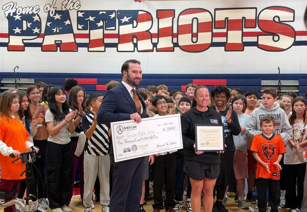 Person in suit holding a big check standing next to teacher holding a certificate in school gym in front of staff and students