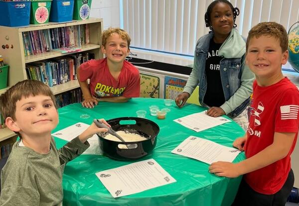 Four children gathered around a green table in a classroom, mixing ingredients in a large black bowl with papers in front of them.