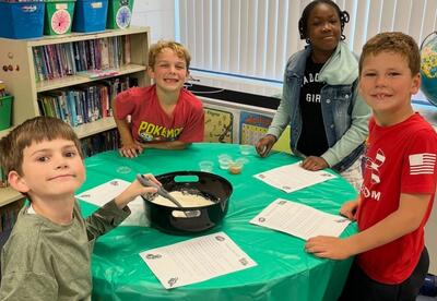 Four children gathered around a green table in a classroom, mixing ingredients in a large black bowl with papers in front of them.