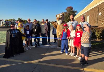 Students in costumes, principal, superintendent and board of education members pose proudly outside of school holding a ribbon to open new playground and blacktop