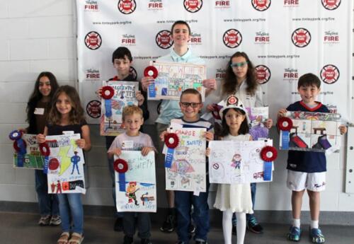 A group of students stands in front of a banner, holding colorful, safety-themed posters and wearing red-and-blue paper badges and ribbons.