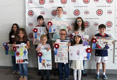 A group of students stands in front of a banner, holding colorful, safety-themed posters and wearing red-and-blue paper badges and ribbons.