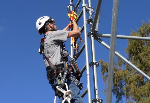 A student wearing a safety harness with clip hooks practices climbing safety on a ladder.