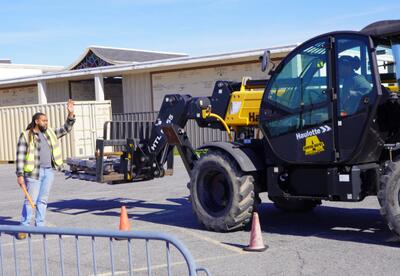 A student wearing a safety vest signals directions to another student operating a telescoping forklift.