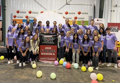 A large group of Pleasant Valley High School students wearing matching purple shirts that read “We Compete So Everyone Can Eat” pose together at the River Bend Food Bank. They’re smiling and holding a banner that says “Student Hunger Drive – 2025 1st Plac