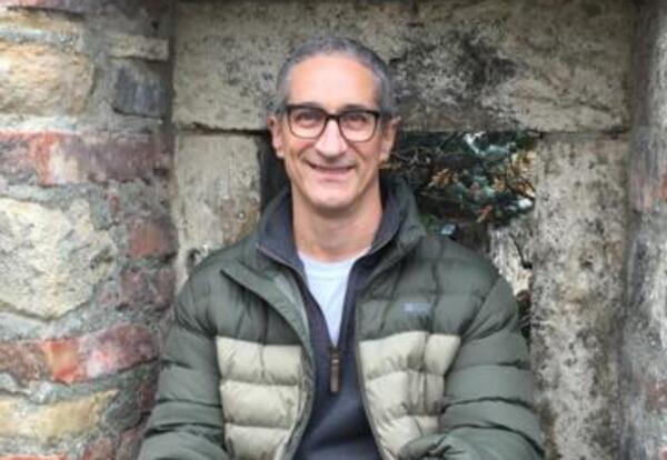 cross-legged in a stone window alcove, wearing glasses, a green puffer jacket, dark jeans, and sneakers, smiling at the camera.