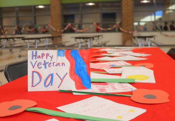 Handmade Veterans Day cards and paper poppy decorations displayed on a red tablecloth in a school cafeteria, with paper chains hanging in the background.