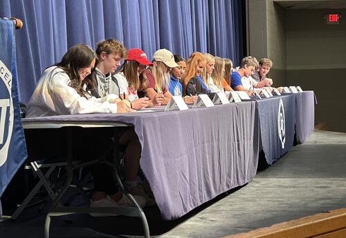 A row of high school student-athletes sit at a long table on stage, signing their National Letters of Intent. Nameplates are lined up in front of them, and a blue curtain hangs behind. A Pleasant Valley banner is visible near the podium, where a speaker a