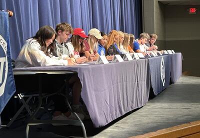 A row of high school student-athletes sit at a long table on stage, signing their National Letters of Intent. Nameplates are lined up in front of them, and a blue curtain hangs behind. A Pleasant Valley banner is visible near the podium, where a speaker a