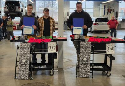 Two Pleasant Valley High School automotive students stand inside a school auto lab, each posing behind a rolling Snap-on tool cart labeled “1st Place.” Both students are holding certificates and standing beside their instructor. The tool carts are filled 