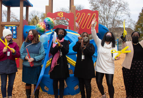 Six people cut a ribbon in front of playground equipment