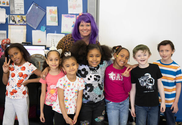 Teacher wearing purple wig stands with seven kids with funny hats and funny hairstyles