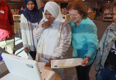 A student putting a mock ballot into a counting machine with the help of a volunteer from the League of Women Voters