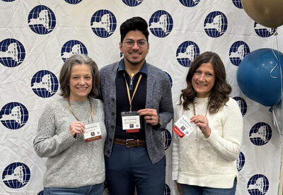 Three teachers wearing their presenter badges stand in front of a conference banner