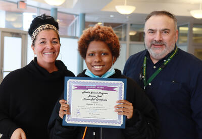 Smiling student holds honor roll certificate with principal and assistant principal standing on either side