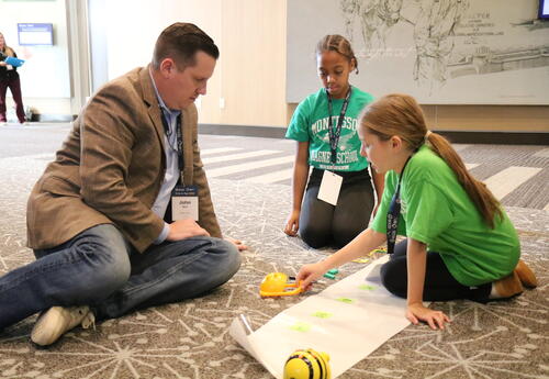 A tech coach works with two students on a math robot project.