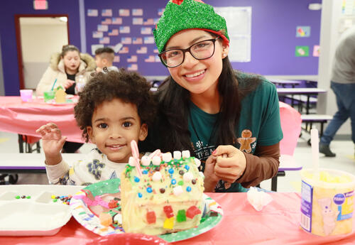 Teacher in a holiday hat helps student decorate gingerbread house