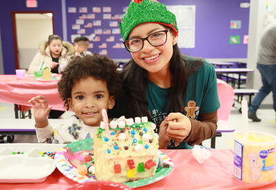 Teacher in a holiday hat helps student decorate gingerbread house