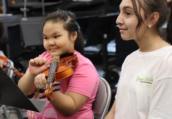 A student learns to play a violin with help from another student.