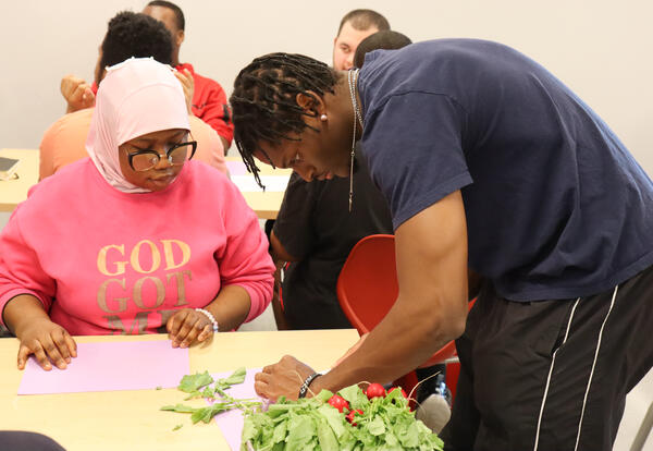 Student observing a volunteer from The Sylvia Center showing how to cut radishes