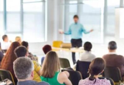 A blur image of a man standing in front of people sitting at a meeting