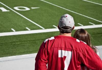 Two people with backs turned standing looking at a football field, red jerseys on.
