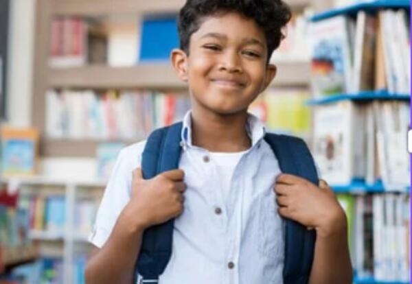 a kid standing with his backpack on and smiling in a library