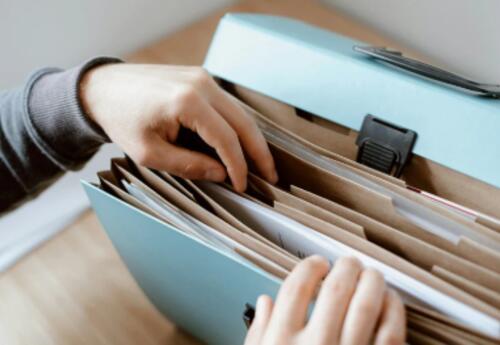 A close-up image shows a person organizing paper documents inside an expanding file organizer with multiple labeled sections. The photo represents gathering and organizing important paperwork needed for student registration, such as proof of residency, id