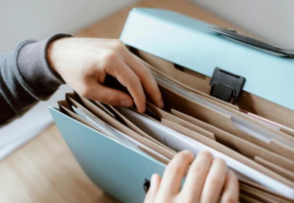 A close-up image shows a person organizing paper documents inside an expanding file organizer with multiple labeled sections. The photo represents gathering and organizing important paperwork needed for student registration, such as proof of residency, id