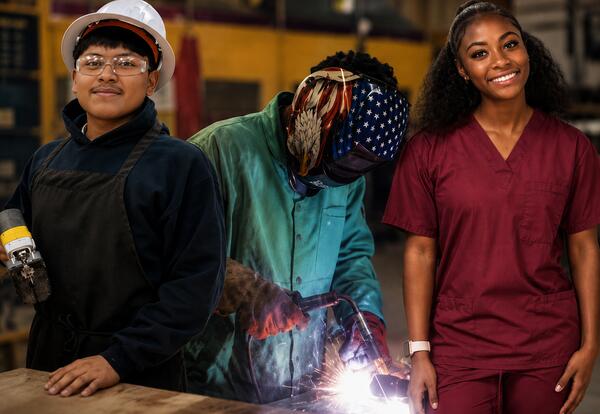 Three Laurel High School students represent Career and Technical Education (CTE) pathways. A student wearing safety glasses, a hard hat, and a work apron stands on the left, representing skilled trades. In the center, a student welds metal while wearing p